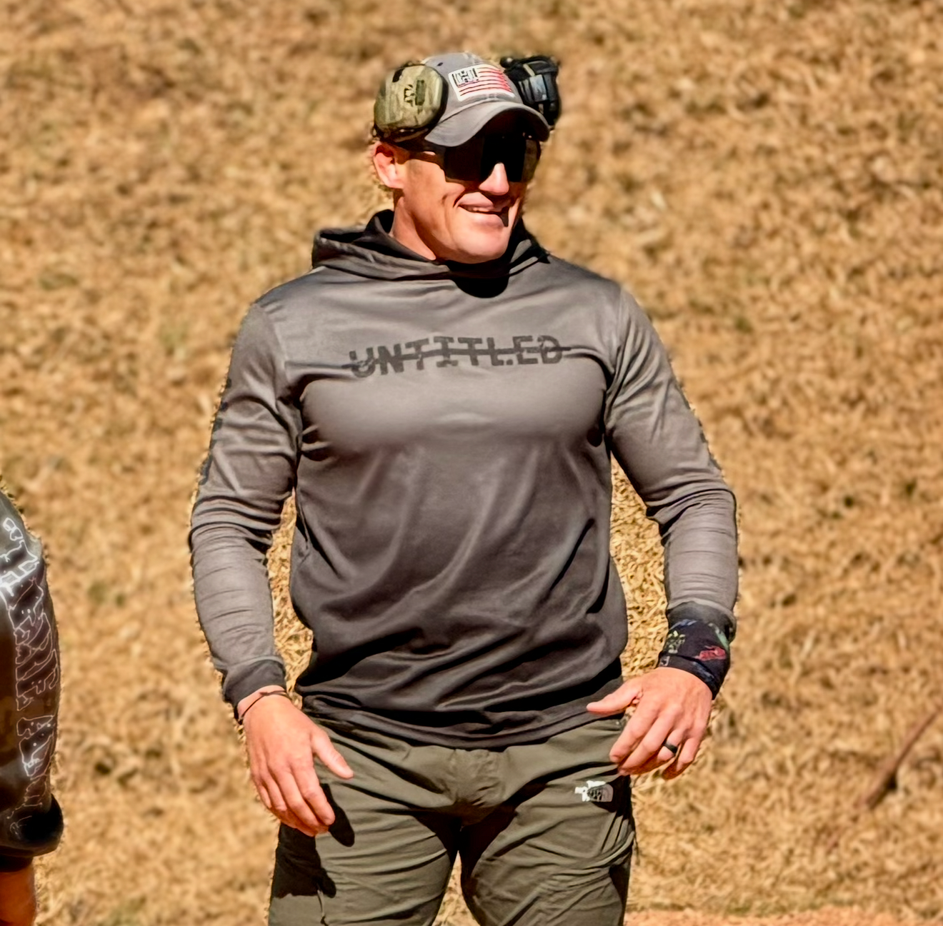 A man wearing a dark gray long-sleeve hooded shirt with 'UNTITLED' printed across the chest, olive green pants, and a camouflage cap, standing outdoors on dry grass.